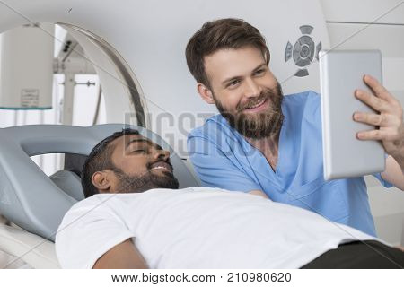 Radiologist Showing Digital Tablet To Patient Lying On CT Scanne