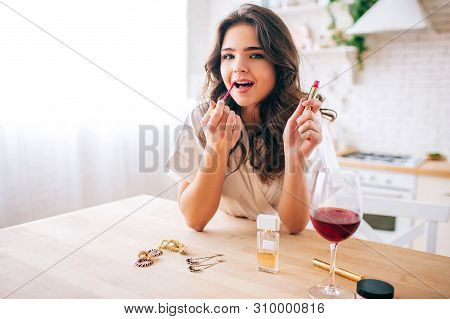 Young Woman With Dark Hair Standing In Kitchen And Putting Make Up On. Glss Of Red Wine On Table. Al