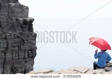 Woman With A Red Umbrella Sits On A Cliff
