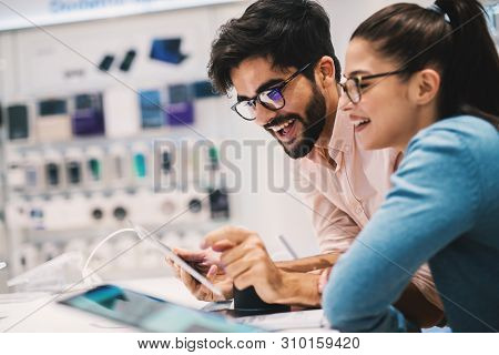 Pretty Young Hipster Couple Deciding Which Mobile Phone To Buy While Shopping In The Mobile Shop.