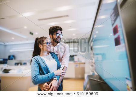 Joyful Young Attractive Hipster Couple Checking Out Plasma Tvs In The Electronic Store.
