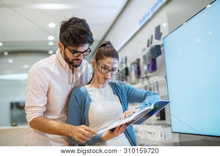Indecisive Young Attractive Couple Shopping In The Electronic Shop Looking Into A Catalogue Of Produ