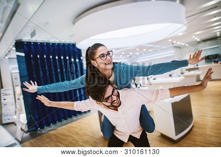 Beautiful Young Modern Couple Having Fun While Shopping In The Electronic Store With A Piggyback Rid