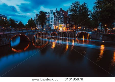 Bridge Illumination And Boat Light Trails In Evening Amsterdam With Reflection In Herengracht Canal.