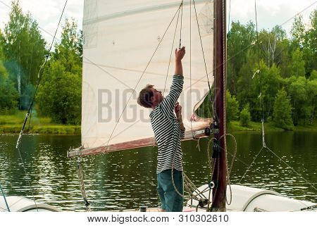 Young Sailor Raises A Sail On A Small Sailing Catamaran Moored In A Wooded Bay