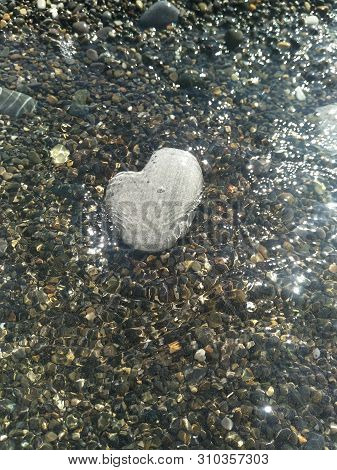 large rounded light grey stone in shape of heart lies under the water near the shore, a running wave, glare on the water, beach pebbles of small colored stones, festive dreamy mood, relax atmosphere, there is free space for writing, copispace, Valentine's