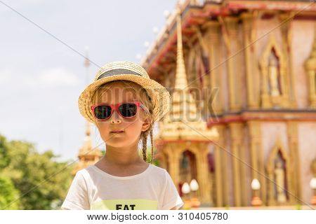 Stylish Little Girl In Straw Hat And Sunglasses Standing Outdoor Near Wat Chalong Temple In Phuket, 
