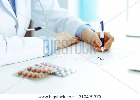 Female Doctor Filling Up Prescription Form While Sitting At The Desk In Hospital. Close-up Of Medica
