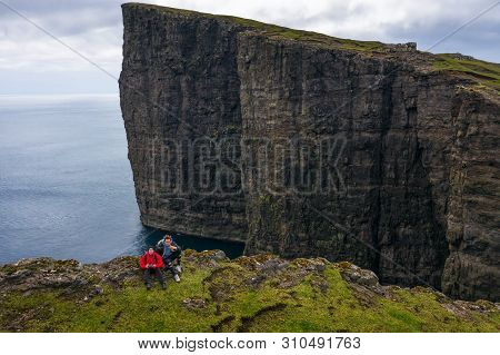 Two Hikers Sitting On The Edge Of A Cliff With The Famous Traelanipan Cliff In The Background. This 