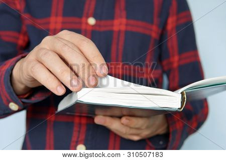 Casually Dressed Young Man Holding An Open Book Flicking Through The Pages. Young Learner Studies Te