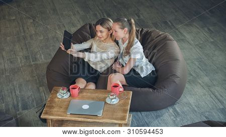 Two Women Take A Photo Yourselves With Digital Tablet In Cafe. Sitting On The Sofa Eating Desserts A