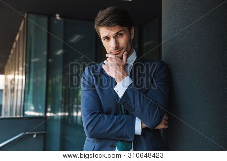 Photo of a thinking young unshaved handsome business man outdoors at the street near business center posing raise one eyebrow.