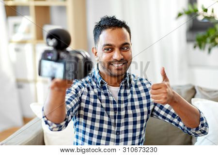 blogging, videoblog and people concept - smiling indian male video blogger with camera videoblogging and showing thumbs up at home