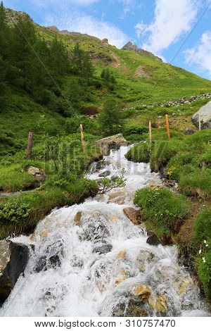 Predatory Mountain River In The Mountains Of The Taur Alps, Green Pasture With Blue Sky, Austria