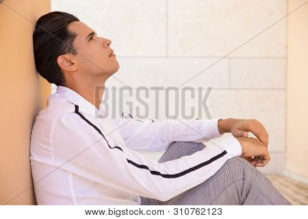 Pensive Latin Man Sitting And Reclining On Wall Outdoors. Handsome Young Man Wearing Shirt And Sitti