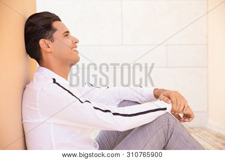 Smiling Latin Man Sitting And Reclining On Wall Outdoors. Handsome Young Man Wearing Shirt And Sitti