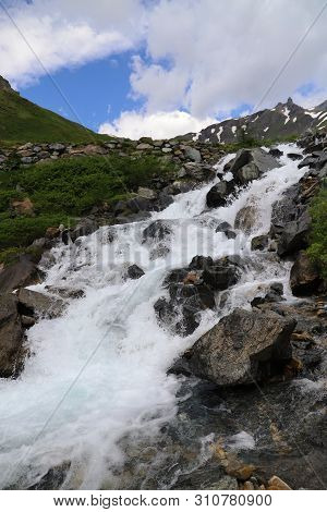 Predatory Mountain River In The Mountains Of The Taur Alps, Green Pasture With Blue Sky, Austria
