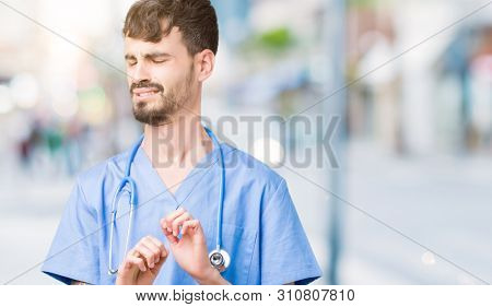 Young handsome nurse man wearing surgeon uniform over isolated background disgusted expression, displeased and fearful doing disgust face because aversion reaction. With hands raised. 