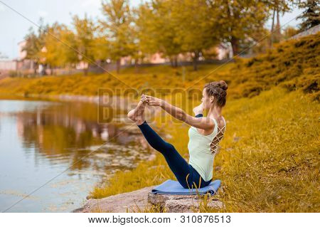 Slender Young Brunette Yogi Performs Challenging Yoga Exercises On The Grass In Autumn Against The B