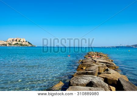 CALVI, FRANCE - SEPTEMBER 21, 2018: Some people diving on the sea and sunbathing in a breakwater at the Plage de Calvi beach, in Calvi, Corsica, France, with its famous citadel in the background