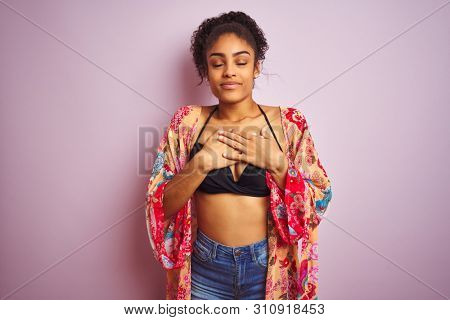 American woman on holiday wearing bikini and colorful dress over isolated pink background smiling with hands on chest with closed eyes and grateful gesture on face. Health concept.