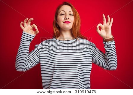 Young redhead woman wearing strapes navy shirt standing over red isolated background relax and smiling with eyes closed doing meditation gesture with fingers. Yoga concept.