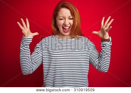 Young redhead woman wearing strapes navy shirt standing over red isolated background celebrating mad and crazy for success with arms raised and closed eyes screaming excited. Winner concept