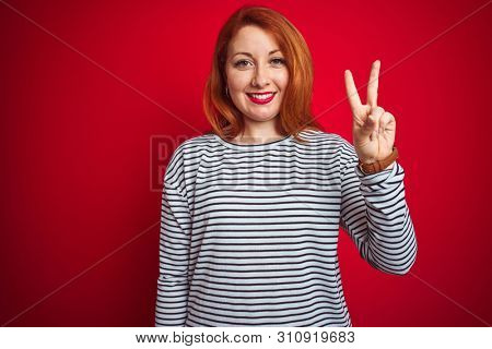 Young redhead woman wearing strapes navy shirt standing over red isolated background showing and pointing up with fingers number two while smiling confident and happy.
