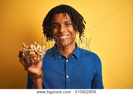 Afro american man with dreadlocks eating peanuts over isolated yellow background with a happy face standing and smiling with a confident smile showing teeth
