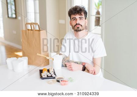 Young man eating asian sushi from home delivery with serious expression on face. Simple and natural looking at the camera.