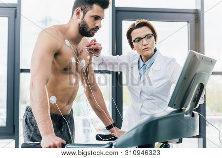 Beautiful Doctor Putting Electrodes On Shirtless Sportsman During Endurance Test