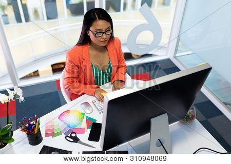 High angle view of pretty young Asian female graphic designer working on computer at desk in office. This is a casual creative start-up business office for a diverse team
