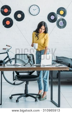 Pretty Radio Host Using Laptop And Holding Coffee Cup While Standing At Wokplace In Studio