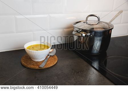 Kitchen Tabletop With Aluminum Cooking Pot On The Vitroceramic, And A Bowl Of Chicken Soup On The Si