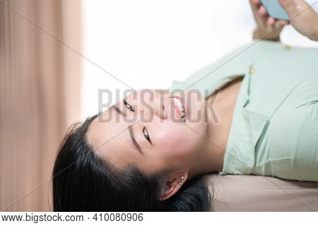 Beautiful Asian Woman Is Lying Down And Playing Mobile On The Bed In Her Bedroom With A Pastel Green