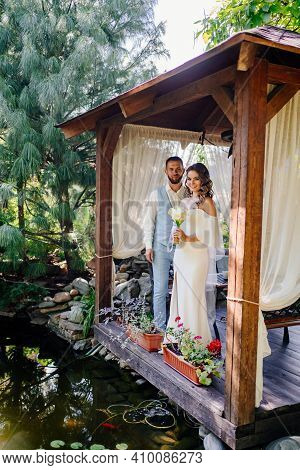 Groom In Blue Suit And Bride With Long Curls With Bouquet In A Gazebo