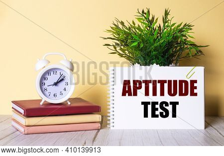 Aptitude Test Is Written In A Notebook Next To A Green Plant And A White Alarm Clock, Which Stands O
