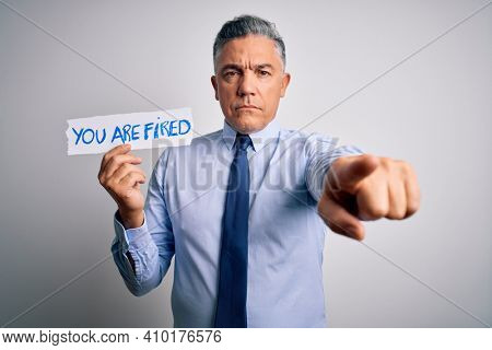 Middle age grey-haired man fired with problems holding paper with you are fired message pointing with finger to the camera and to you, hand sign, positive and confident gesture from the front