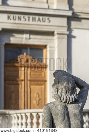 Chur, Switzerland - February 19, 2021: A Sculpture Of A Man Looking Towards The Old Building Of The 