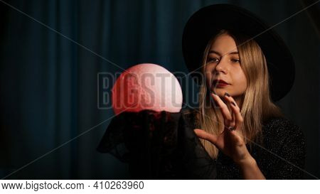 A Young Woman Fortune Teller In A Hat Is Holding A Magic Ball. Beautiful Psychic Woman Is Guessing W