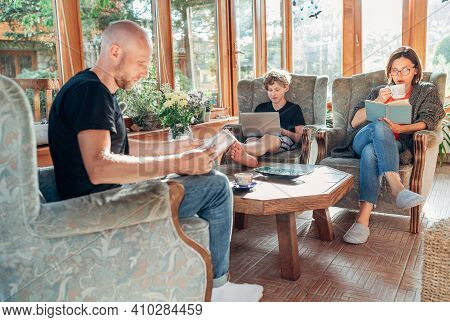 Mother, Father And Son Family Sitting Together In Sunroom In Cozy Armchairs And Reading Books, Using
