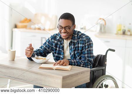 Positive Black Impaired Guy In Wheelchair Having Breakfast And Reading Interesting Book At Kitchen. 