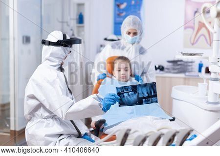 Child And Mother In Ppe Suit Listening Dentist Talking About Dental Procedure. Stomatolog In Protect