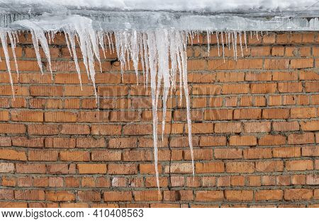 Long Icicles Of Ice Hang From The Roof Of A Red-brick House In Early Spring. Thaw And The Danger Of 