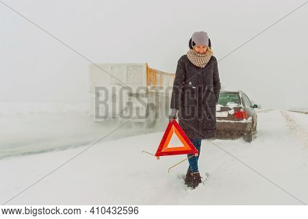 Young Girl In A Jacket, Hat, Scarf, Shoes Puts A Warning Road Sign In A Triangle On A Snow-covered R