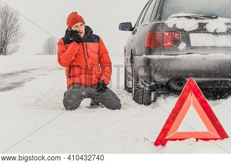 Angry Young Man In An Orange Jacket Is On His Knees Talking On A Mobile Phone On A Snowy Road. He Is