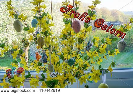 Colored Easter Eggs Standing On A Forsythia Bush In A Vase In Front Of A Window. Blur Background