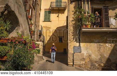 Old Woman On Balcony And  Man In Black Mask During Coronavirus Epidemic In  Medieval Town Of Orte In