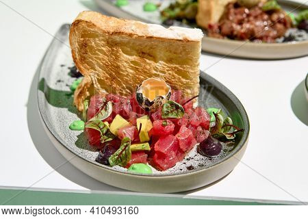 Tuna tartar with quail egg and toast bread. Raw fish tartare restaurant appetizer on white table with green wall. Day sunlight with hard shadow of monstera palm leaves. Summer or spring food concept
