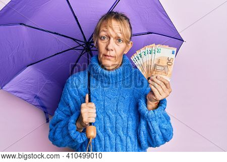 Middle age blonde woman holding umbrella and 50 euros banknotes in shock face, looking skeptical and sarcastic, surprised with open mouth 